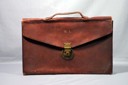 Brown leather briefcase with a rectangular flap featuring a gold metal clasp and a grip on the top. The letters W.L. (Wilfrid Laurier) are inscribed in gold at the front.
