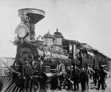 Photo of a locomotive with a funnel, decorated with garlands and Queen Victoria’s coat of arms for her Jubilee. The locomotive pulls several wagons. A group of men alongside the train.